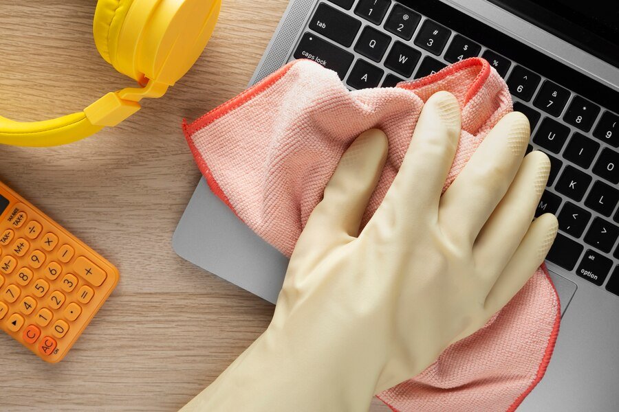 Person cleaning a laptop with a cloth, yellow headphones, and an orange calculator nearby