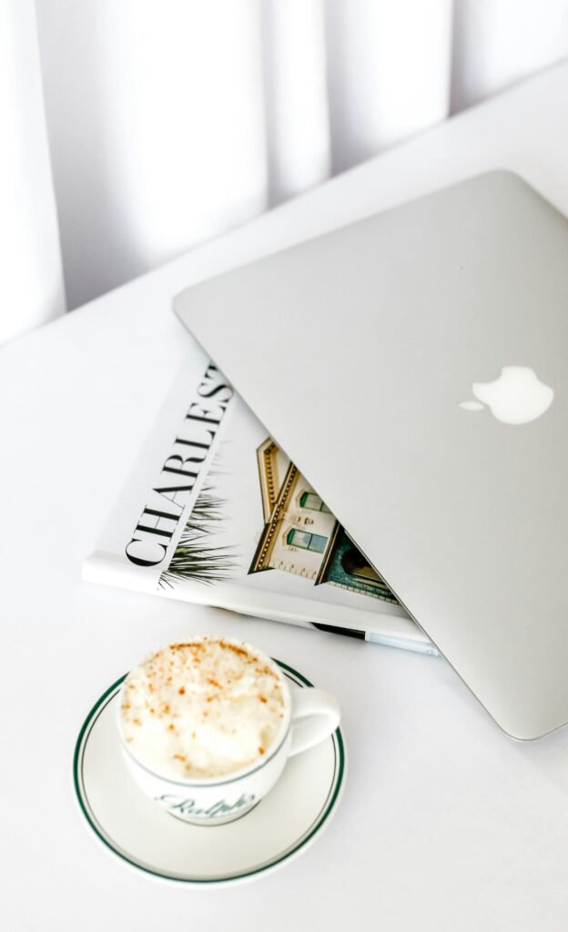 A MacBook, a newspaper, a pen & a cup of tea placed on a white counter top